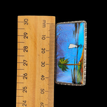 Load image into Gallery viewer, A butterfly wing brooch depicting a sailboat with a palm tree in the foreground, set in silver, next to a ruler on a black background