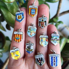 Load image into Gallery viewer, Enamelled silver shield charms depicting various towns, cities and countries, held in a hand