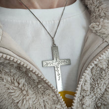 Load image into Gallery viewer, A silver cross pendant engraved with a bird and vines, with a curb link chain, modelled by a woman wearing a white t-shirt and fluffy jacket
