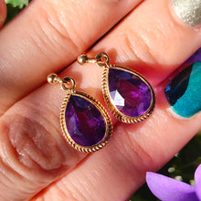 Load image into Gallery viewer, A pair of pear shaped amethyst drop earrings in yellow gold rope edge mounts, held in a hand