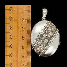 Load image into Gallery viewer, An oval silver locket pendant with acorn and oak leaf detailing next to a wooden ruler, on a black background