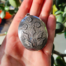 Load image into Gallery viewer, A silver oval locket with flower details, held in a hand, with leaves in the background