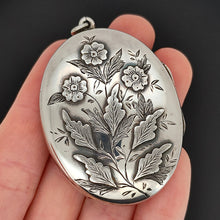 Load image into Gallery viewer, A silver oval locket with flower details, held in a hand, on a black background
