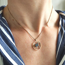 Load image into Gallery viewer, A circular cutout pendant featuring a wading bird with a blue paste stone body, in yellow gold, with a chain and modelled by a woman wearing a stripey shirt