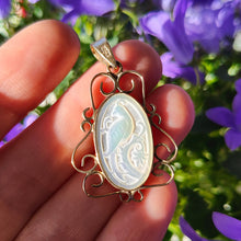Load image into Gallery viewer, An oval mother of pearl and yellow gold pendant with a carved bird, held in a hand, with purple flowers in the background