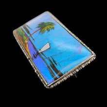 Load image into Gallery viewer, A butterfly wing brooch depicting a sailboat with a palm tree in the foreground, set in silver, on a black background
