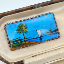 Load image into Gallery viewer, A butterfly wing brooch depicting a sailboat with a palm tree in the foreground, set in silver, in an antique box
