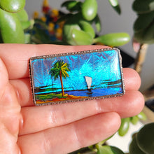 Load image into Gallery viewer, A butterfly wing brooch depicting a sailboat with a palm tree in the foreground, set in silver, held in a hand
