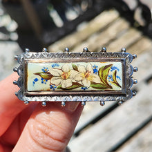 Load image into Gallery viewer, An oblong silver hobnail bead border brooch with an enamelled central plaque displaying flowers, held in a hand
