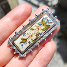 Load image into Gallery viewer, An oblong silver hobnail bead border brooch with an enamelled central plaque displaying flowers, held in a hand
