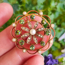 Load image into Gallery viewer, A circular brooch/pendant in yellow gold with peridots and pearls, with a flower to the centre, held in a hand
