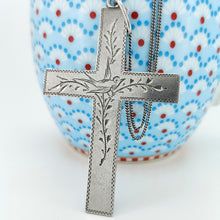 Load image into Gallery viewer, A silver cross pendant engraved with a bird and vines, with a curb link chain, leaning against a blue, red and white jug
