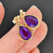 Load image into Gallery viewer, A pair of pear shaped amethyst drop earrings in yellow gold rope edge mounts, held in a hand
