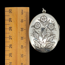 Load image into Gallery viewer, A silver oval locket with flower details next to a wooden ruler, on a black background
