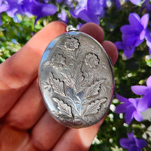 Load image into Gallery viewer, A silver oval locket with flower details, held in a hand, with purple flowers in the background
