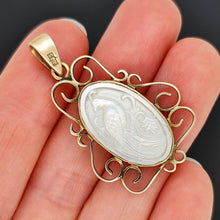 Load image into Gallery viewer, An oval yellow gold and mother of pearl pendant carved with a bird, held in a hand
