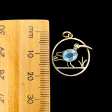 Load image into Gallery viewer, A circular cutout pendant featuring a wading bird with a blue paste stone body, in yellow gold, next to a ruler on a black background
