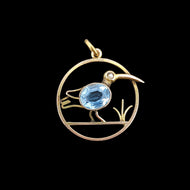 A circular cutout pendant featuring a wading bird with a blue paste stone body, in yellow gold, on a black background