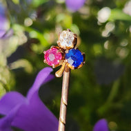 A ruby, sapphire and diamond clover shaped stick tie pin in gold, on a floral background